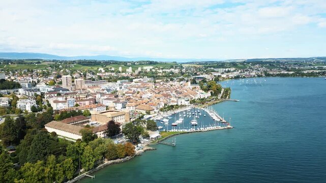 Aerial view of Morges, a city with terracotta rooftops, a marina filled with boats, and green trees bordering the turquoise lake, Morges, Vaud, Switzerland.