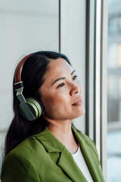 Businesswoman in green jacket listening with headphones by window