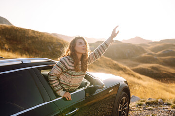 A happy traveler leans out of a car window at sunset. The young woman feels free and enjoys the mountain scenery during her drive. Adventure and transportation concept. Active lifestyle.