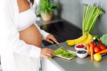 A young pregnant woman chops celery while standing in a modern kitchen. A beautiful expectant...