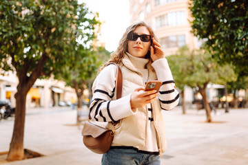A young woman in sunglasses strolls along the sunny streets of the city with her phone. A tourist...