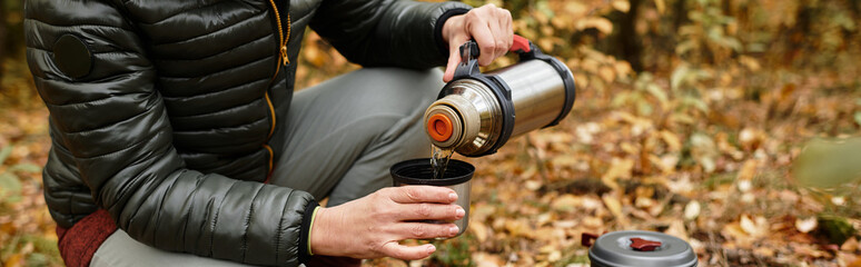 Naklejka premium Senior woman enjoys a warm drink while hiking in a vibrant forest setting on a cool day