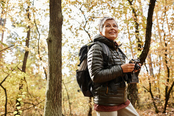 Fototapeta premium Joyful senior woman hiking through a vibrant forest in autumn sunshine with camera in hand