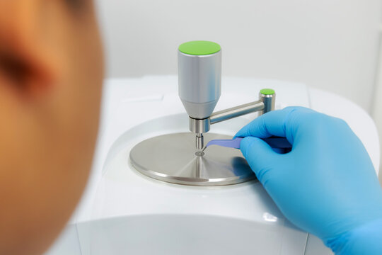 A laboratory technician carefully places a sample to the analysis plate of an FT-IR (Fourier Transform Infrared) spectrometer using tweezers prepared for precise material laboratory technology.