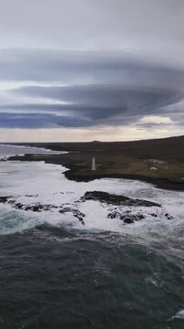 Aerial view of the white Malarrif Lighthouse standing tall against the dramatic coastline with crashing waves and a cloudy sky, Hellnar, Sn&aelig;fellsb&aelig;r, Iceland.