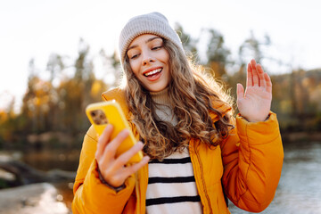 Cute woman in a bright yellow jacket talks on her phone in a sunny forest by a river. A beautiful traveler texts and enjoys the sunset outdoors. Adventure and blogging concept.