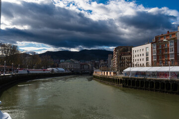 View of the Bilbao river with the Arriaga Theater in the background under a cloudy sky.