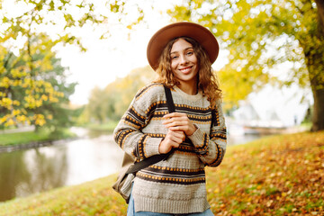 A cheerful woman in a hat and raincoat stands by a river in an autumn park. A young tourist with a backpack enjoys nature and an outdoor walk. Concept of seasonality and tourism. © maxbelchenko