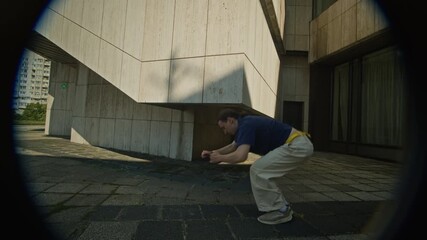 Tilt down iris shot of acrobatic young Caucasian man in urbanwear performing backflip off concrete wall practicing extreme parkour stunts at high rise industrial site on sunny day - Powered by Adobe