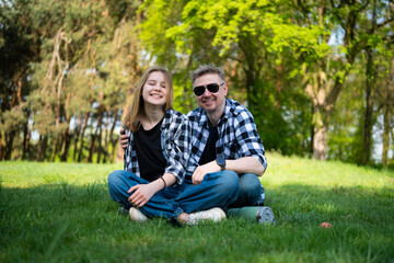 Happy Father And Daughter Sit On The Grass In The Park, Looking Into The Camera And Enjoying The Outdoors
