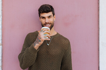 Handsome man drinking coffee from a disposable cup smiling near pink wall