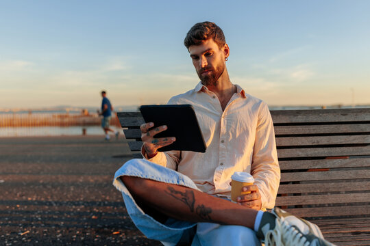 Handsome young man using tablet and drinking coffee outdoors at sunset