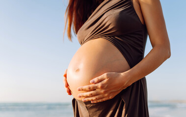 A pregnant woman stands on the beach, gently touching her belly. A close-up of a woman expecting a baby enjoying the sunset on the seashore. Concept of nature and relaxation.