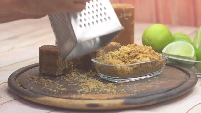 Close-up shot of a person's hand grating a block of brown panela on a rustic wooden board.