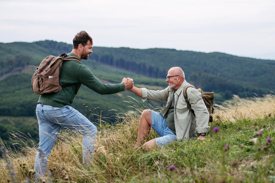 Adult son helping older father to stand up. Hiking togeter in nature, resting on meadow. - Powered by Adobe