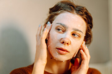 Fototapeta premium Cute woman wearing fabric face mask looking at mirror in cozy room. Beautiful woman performing indoor care procedures, touching face while looking at reflection. Cosmetology and care concept.