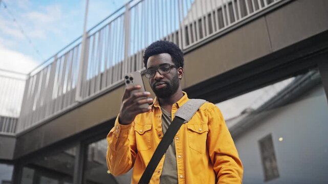 Handsome African American man with beard wearing glasses holding smartphone outdoors. Male with shoulder bag standing near modern building, smiling slightly while reading screen outside.