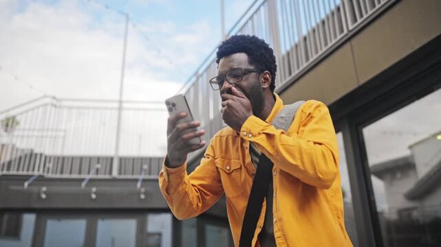 Handsome African American man with beard wearing glasses holding smartphone outdoors, looking surprised. Male reacting emotionally to good news or winning while standing near modern building outside.
