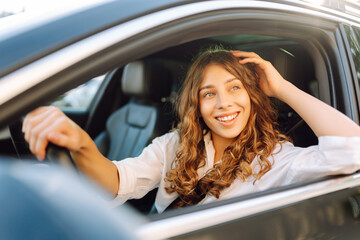 Portrait of smiling woman sitting behind the wheel of car looking out the window. Woman driver in modern car enjoying sunny day. Transport, taxi concept. Lifestyle.