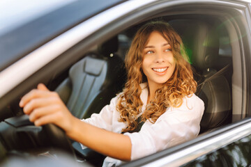 Portrait of smiling woman sitting behind the wheel of car looking out the window. Woman driver in modern car enjoying sunny day. Transport, taxi concept. Lifestyle.