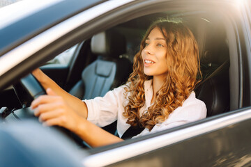 Portrait of smiling woman sitting behind the wheel of car looking out the window. Woman driver in modern car enjoying sunny day. Transport, taxi concept. Lifestyle.