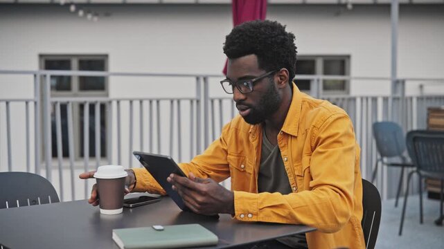 Handsome African American man with beard using digital tablet outdoors at table. Male in glasses concentrating on screen with coffee cup and notebook beside, working or studying outside.