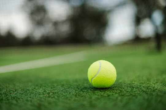 A vibrant yellow tennis ball rests on a green artificial grass court on overcast day
