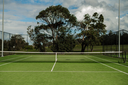 Empty tennis court features green grass and trees surrounding the area, under cloudy skies.