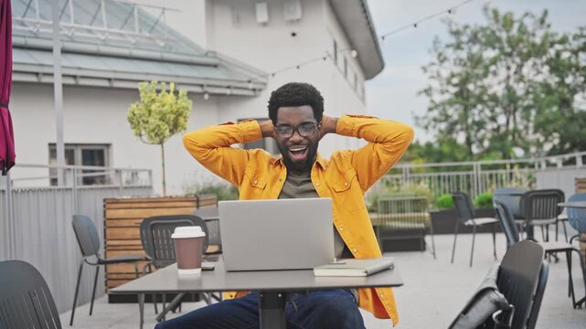 Handsome African American man raising hands in surprise while working on laptop outdoors. Male sitting at table with coffee cup and notebook, reacting emotionally outside to good news or victory.