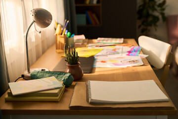 Empty wooden desk with open sketchbook, stack of notebooks, potted plant, desk lamp and scattered colorful drawings suggesting creative workspace for child or teenager