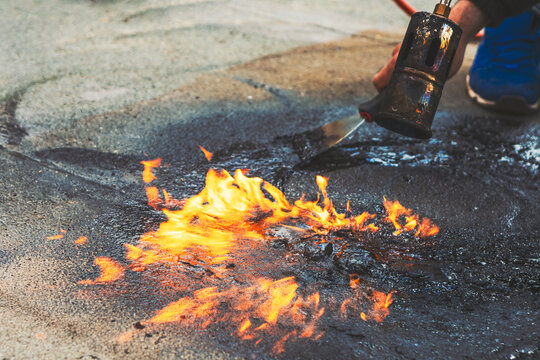 Worker is using a blowtorch to repair a flat rooftop. Flame heats a black, tar substance, causing it to melt and adhere. Process of sealing or repairing asphalt with heat and flame