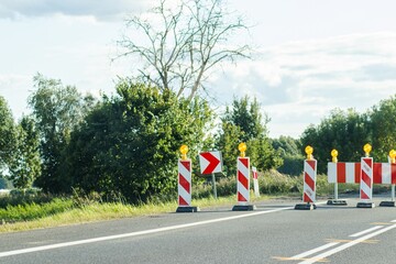 Road construction scene featuring traffic barriers and warning signs, surrounded by lush greenery and a clear sky, indicating ongoing roadwork and safety measures