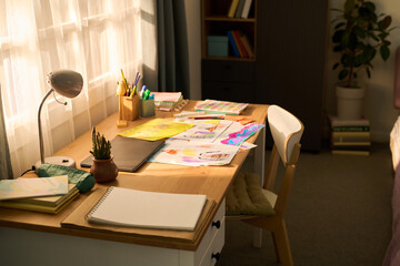 No people view of colorful drawings on desk with sketchbook, colored pencils, and scattered artwork, sunlight streaming through window, home study area with books and plant visible