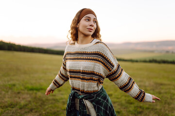 Young woman in a sweater standing on a green hill in the rays of sunset. Traveler enjoying nature and mountain landscape. Nature, recreation concept.
