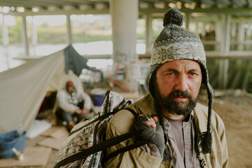 Obraz premium Portrait of middle aged Caucasian man with beard carrying backpack and wearing knit hat standing outdoors under bridge with tent, and another middle aged Black man sitting in background