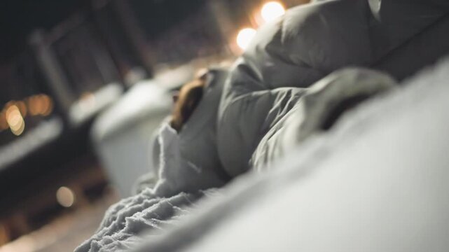 Side view of young girl in winter jacket lying on snow-covered ground swinging arms gently while surrounded by soft bokeh lights and snowy atmosphere in park at night, creating serene