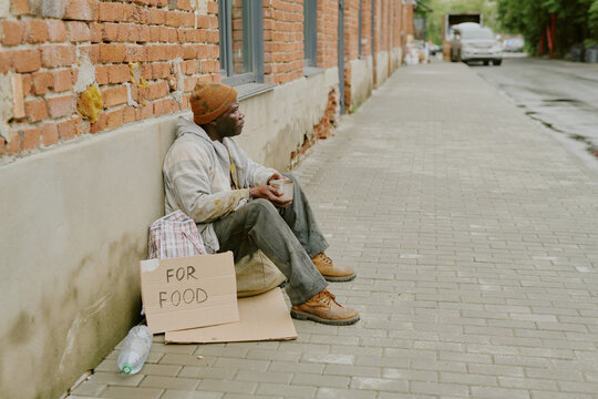 Black middle aged man sitting on sidewalk leaning against brick wall holding cup with cardboard sign reading for food beside him, appearing homeless and seeking assistance