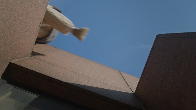 Low angle rotating slowmo of acrobatic young Caucasian man in urbanwear jumping across gap practicing parkour stunts in urban environment on sunny day - Powered by Adobe