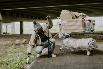 Middle aged Caucasian man wearing winter hat bending down with walking stick, picking up bottle...