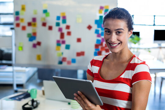 Mid-20s woman smiling and holding tablet in office by whiteboard with sticky notes, copy space