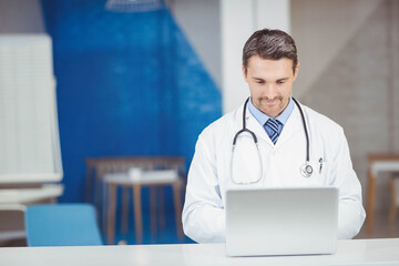 Doctor working on silver laptop at white desk in clinic reception with blue chairs and stethoscope