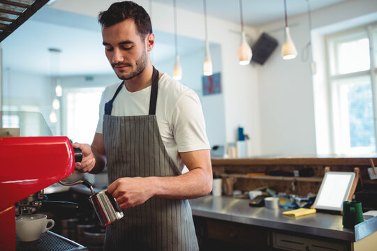 Male barista steaming milk with metal pitcher behind espresso machine at coffee shop counter