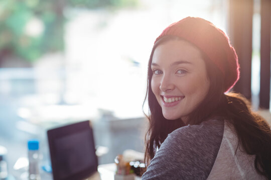 Woman wearing red beanie sitting by sunlit cafe window using laptop beside water bottle, copy space