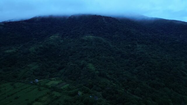 A nearer sky-high shot of the forested Mounta Banahaw veild in low clouds, with dense trees, small fields, and scattered homes during dusk in Quezon Province Philippines