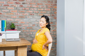 Asian pregnant woman pressing back and sitting at home office desk with laptop, folders, copy space