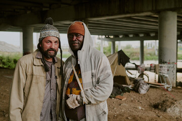 Caucasian middle aged man and Black middle aged man standing together under bridge, smiling at camera, makeshift shelter and shopping cart visible in background, outdoor urban setting