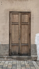 A double wooden door on a beige plaster wall. A worn surface, simple architecture, and stone flooring create a nostalgic street texture.