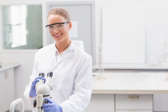 Female scientist wearing lab coat, goggles and gloves, adjusting microscope focus at bench