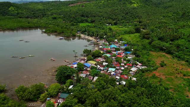 A rotating bird's eye view shot captures clustered shoreline homes nestled in lush greenery, with boats docked nearby and the calm coastal water near Mauban Port, Quezon Province Philippines