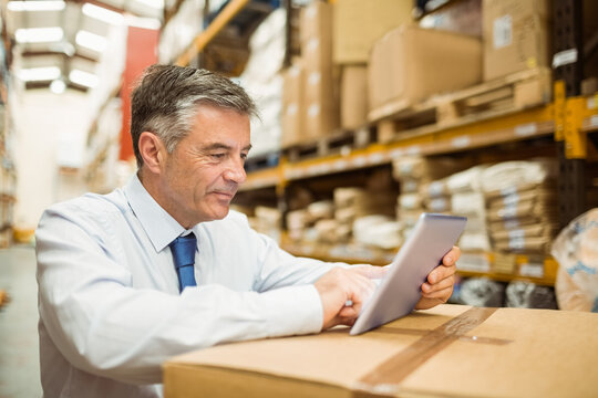 Senior white male manager in business attire standing in warehouse aisle examining tablet on box - Powered by Adobe